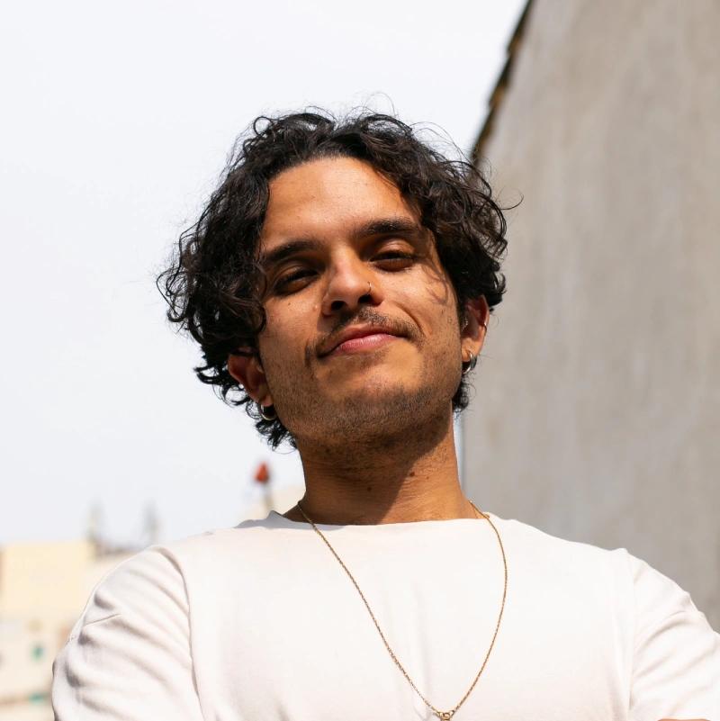 Homem jovem de cabelo castanho-escuro e ondulado, com barba rala e expressão confiante. Ele usa camiseta clara, colar fino dourado, brinco e piercing no nariz. A foto é tirada ao ar livre, com luz natural e fundo urbano desfocado.