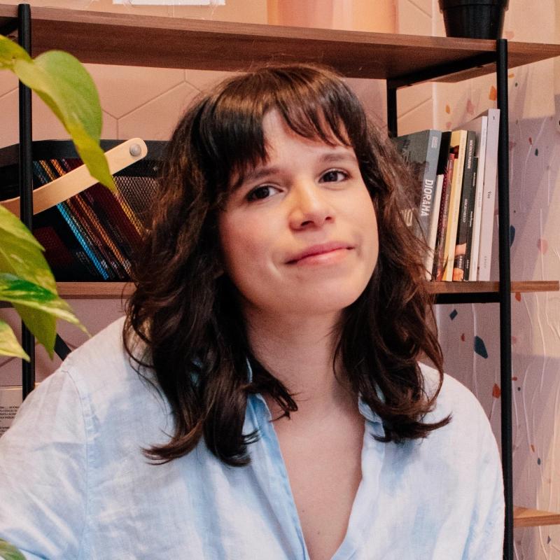 Mulher jovem de cabelos escuros e ondulados, com franja, usando camisa clara, sorrindo suavemente, em ambiente interno com prateleira de livros ao fundo.